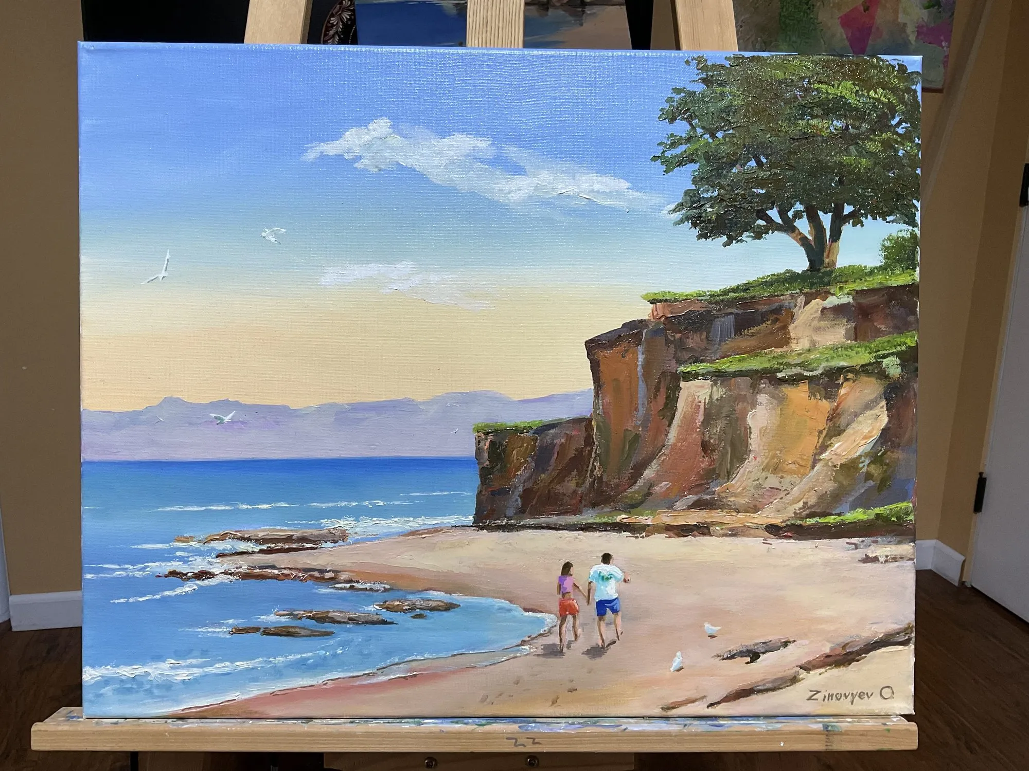 Painting “Study: Rocks” by Oleksandr Zinovyev: A couple walking on a beach below a red cliff with a large tree; photographed on the easel.