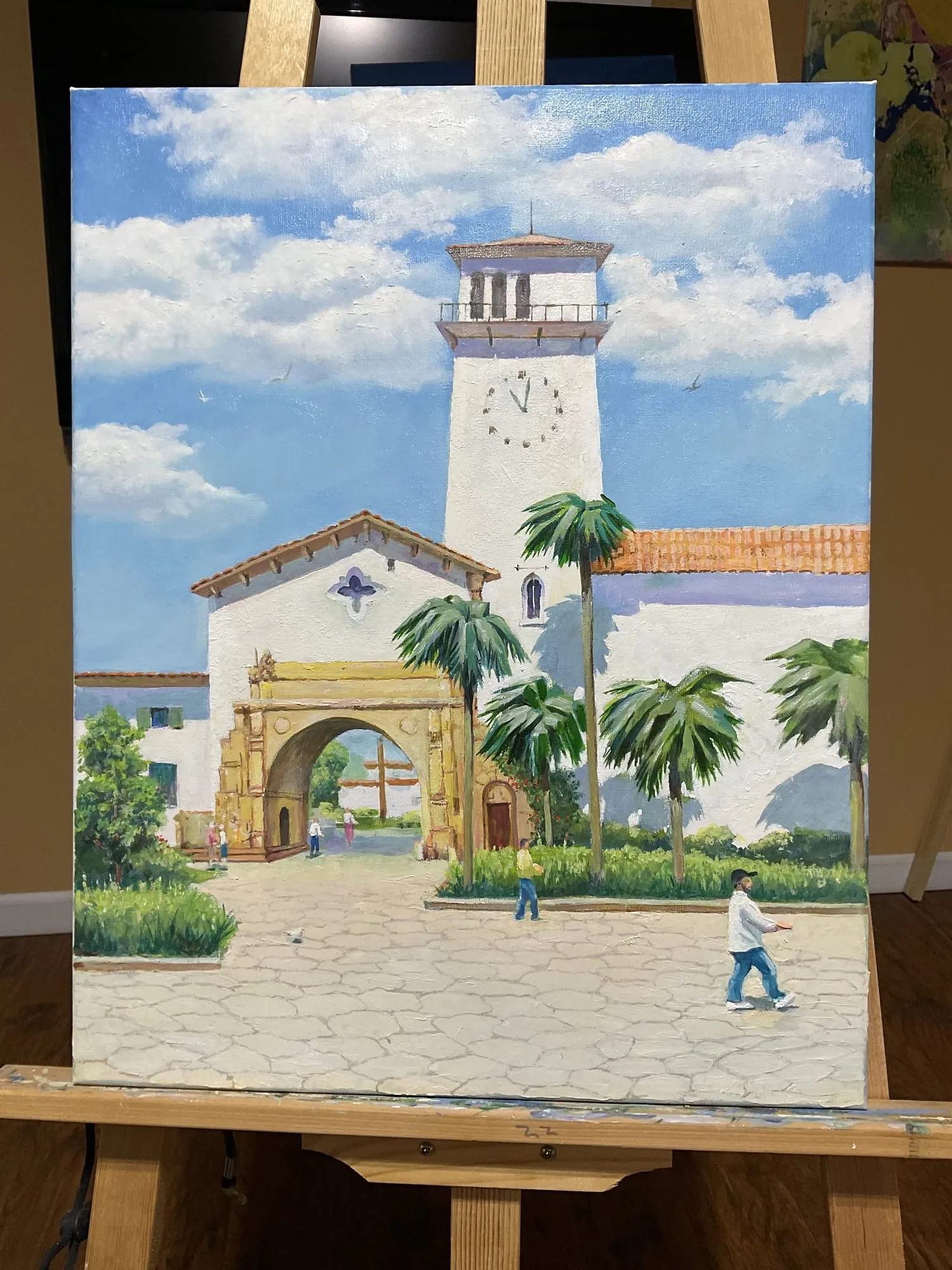 Painting “Study: The Mission, Santa Barbara” by Oleksandr Zinovyev: A white clock tower and arched gateway with palm trees and clouds in Santa Barbara; photographed on the easel.