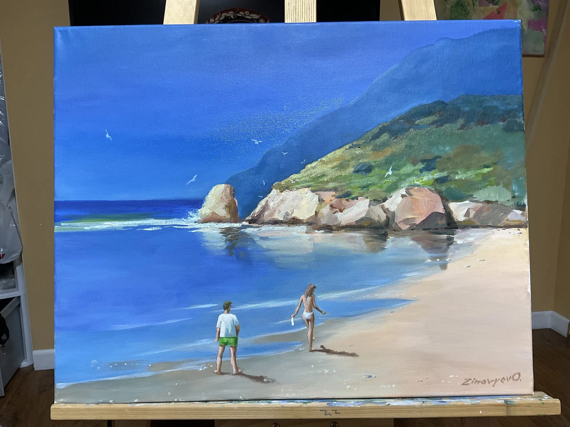 Painting “Study by the Ocean I” by Oleksandr Zinovyev: A turquoise bay with a rocky headland and two people walking along the sand; photographed on the easel.