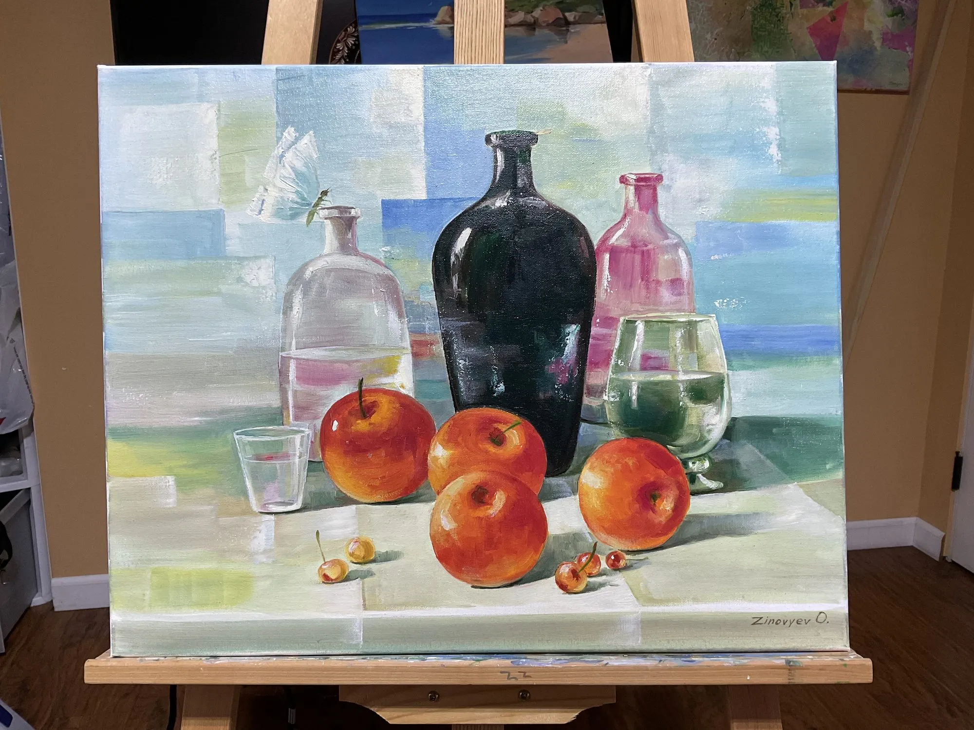 Painting “Still Life by Mood” by Oleksandr Zinovyev: A dark bottle, glass bottles and red apples on a pale table; photographed on the easel.