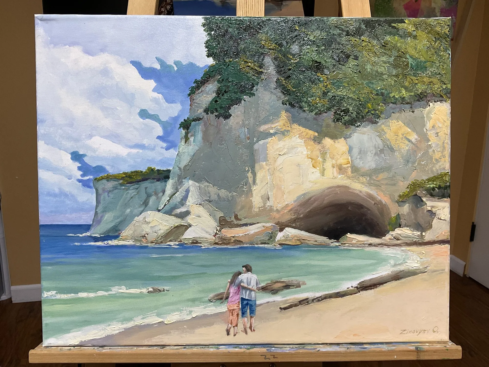 Painting “Walk to the Grotto, Santa Barbara” by Oleksandr Zinovyev: A couple walking towards a large sea cave in a white cliff; photographed on the easel.
