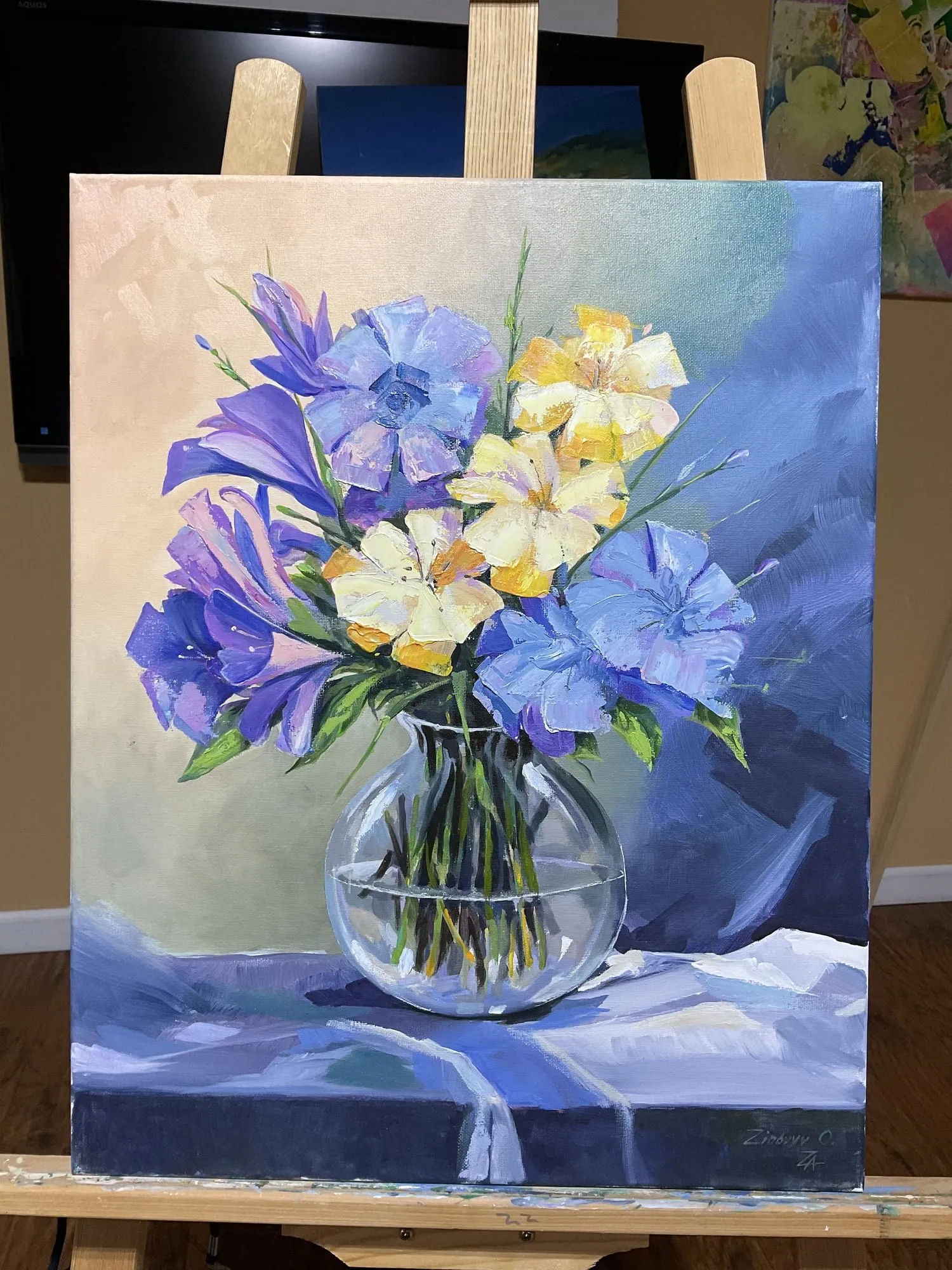 Painting “Blue Flowers on Blue” by Oleksandr Zinovyev: Blue, violet and white flowers in a round glass vase on blue cloth; photographed on the easel.