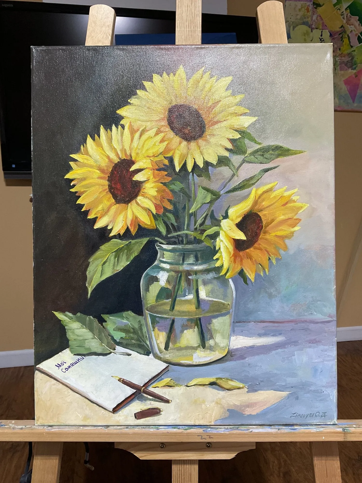 Painting “Sunflowers” by Oleksandr Zinovyev: Sunflowers in a glass jar beside an open notebook and pencils; photographed on the easel.